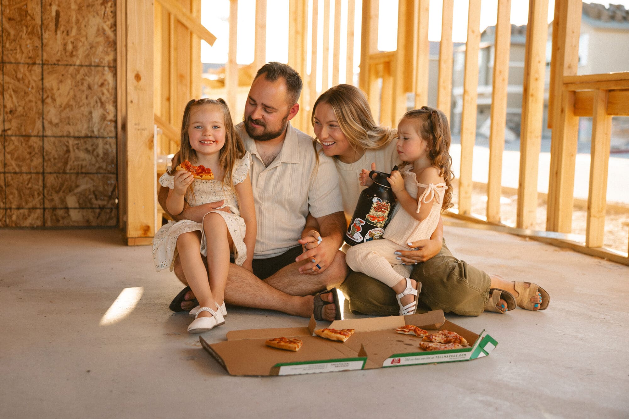 Family of four sitting on the floor of an unfinished wood-framed home, eating pizza together during an in-home family session, with parents smiling at their young children in warm natural light.