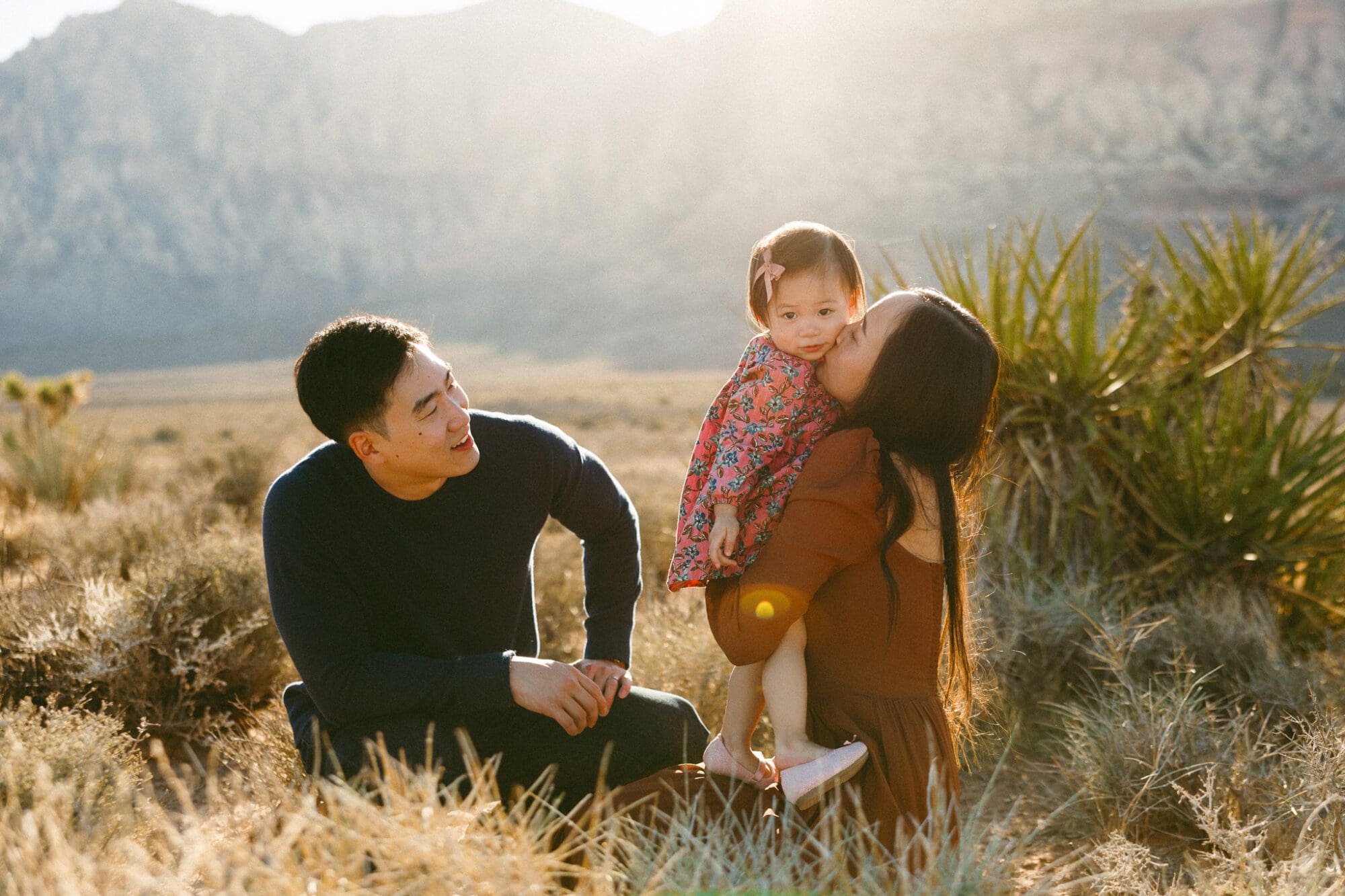family of three, dad kneeling and mom kissing daughters cheek in the golden light of red rock.
