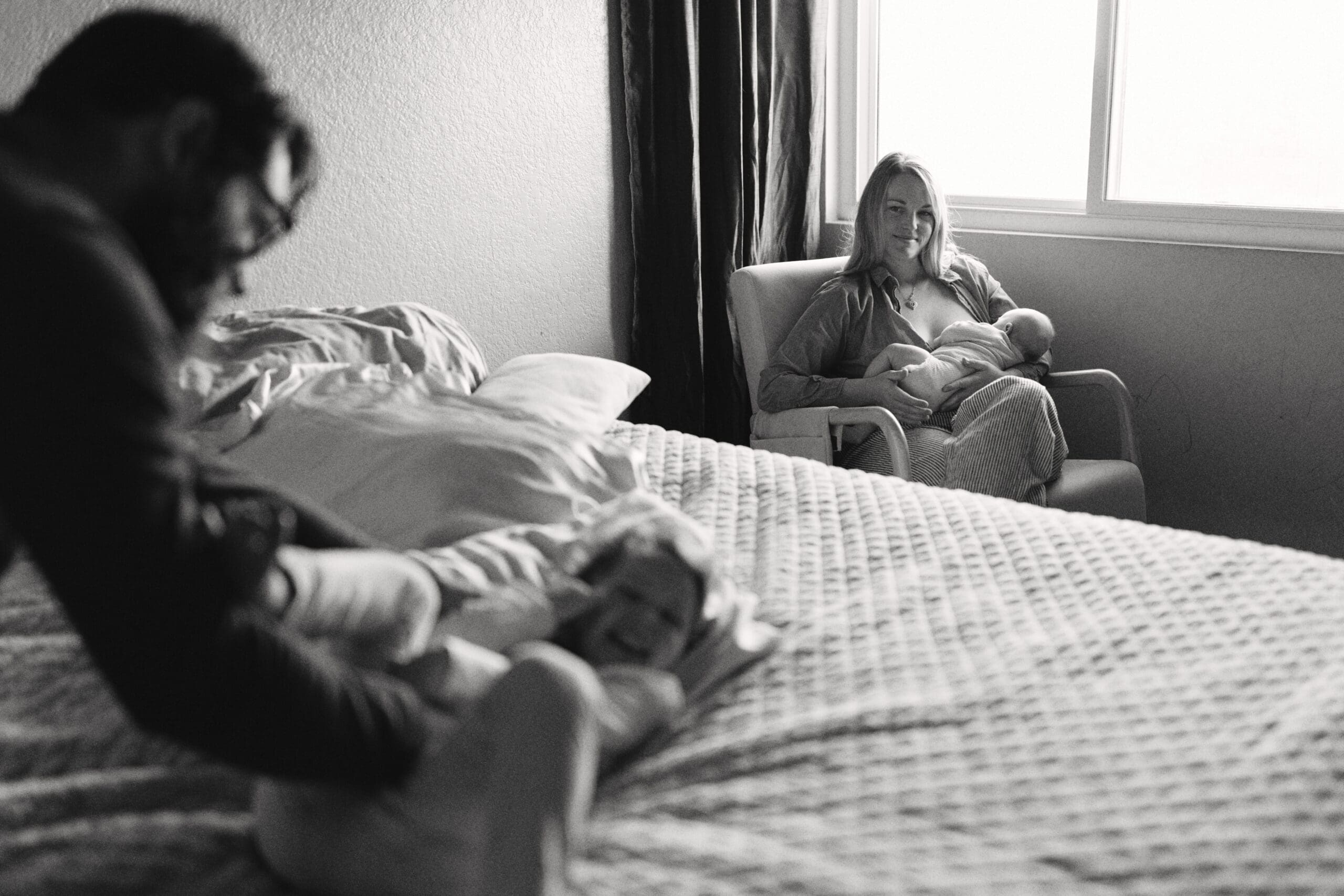 Mom breastfeeding her newborn in a sunlit bedroom chair while dad comforts their toddler on the bed, captured by a Las Vegas family photographer.